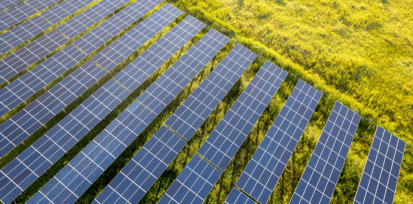 Mehrere Reihen von Solarmodulen verlaufen parallel über eine große, grüne Wiese. Die Module sind rechteckig angeordnet und reflektieren das Tageslicht. Zwischen den Reihen bleibt jeweils ein schmaler Streifen Gras sichtbar.