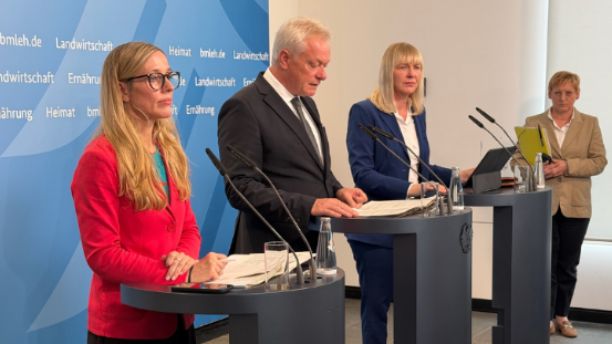 m Vordergrund stehen drei Personen hinter Pulten, die mit Mikrofonen ausgestattet sind. Die mittlere Person trägt einen dunklen Anzug und hält ein Manuskript, während die beiden anderen Personen links und rechts jeweils einen farbigen Blazer tragen. Im Hintergrund befindet sich eine weitere Person mit Tablet. Die Wand hinter den Pulten zeigt das Logo und die Schriftzüge des Bundesministeriums für Ernährung und Landwirtschaft. Die Szene vermittelt den Eindruck einer Pressekonferenz zum Erntebericht, bei der Alois Rainer und Nikola Steinbock im Mittelpunkt stehen.
